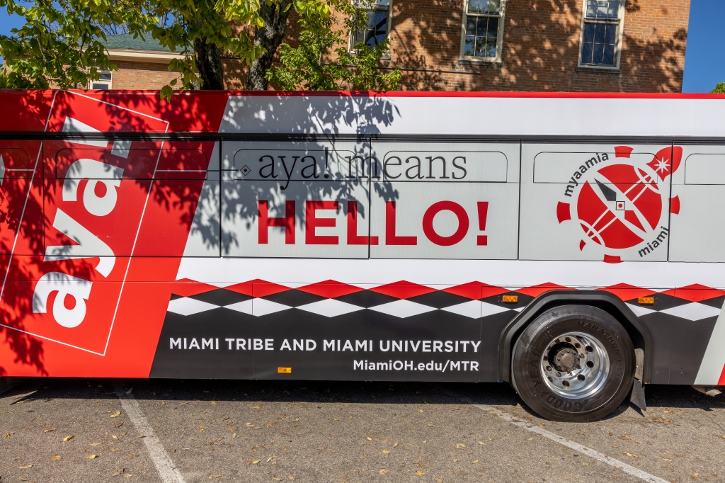 A metro bus with red and black text reading "aya! aya means hello." The bus is decorated with rows of triangles and logo in the shape of a turtle that reads "Myaamia" and "Miami." 