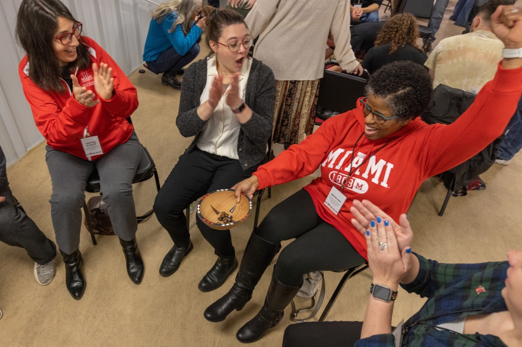 3 guests from Miami University sit in chairs in a circle to play a game involving the tossing of marked beans in a bowl. The person holding the bowl pumps their arm in the air with excitement about their score. 