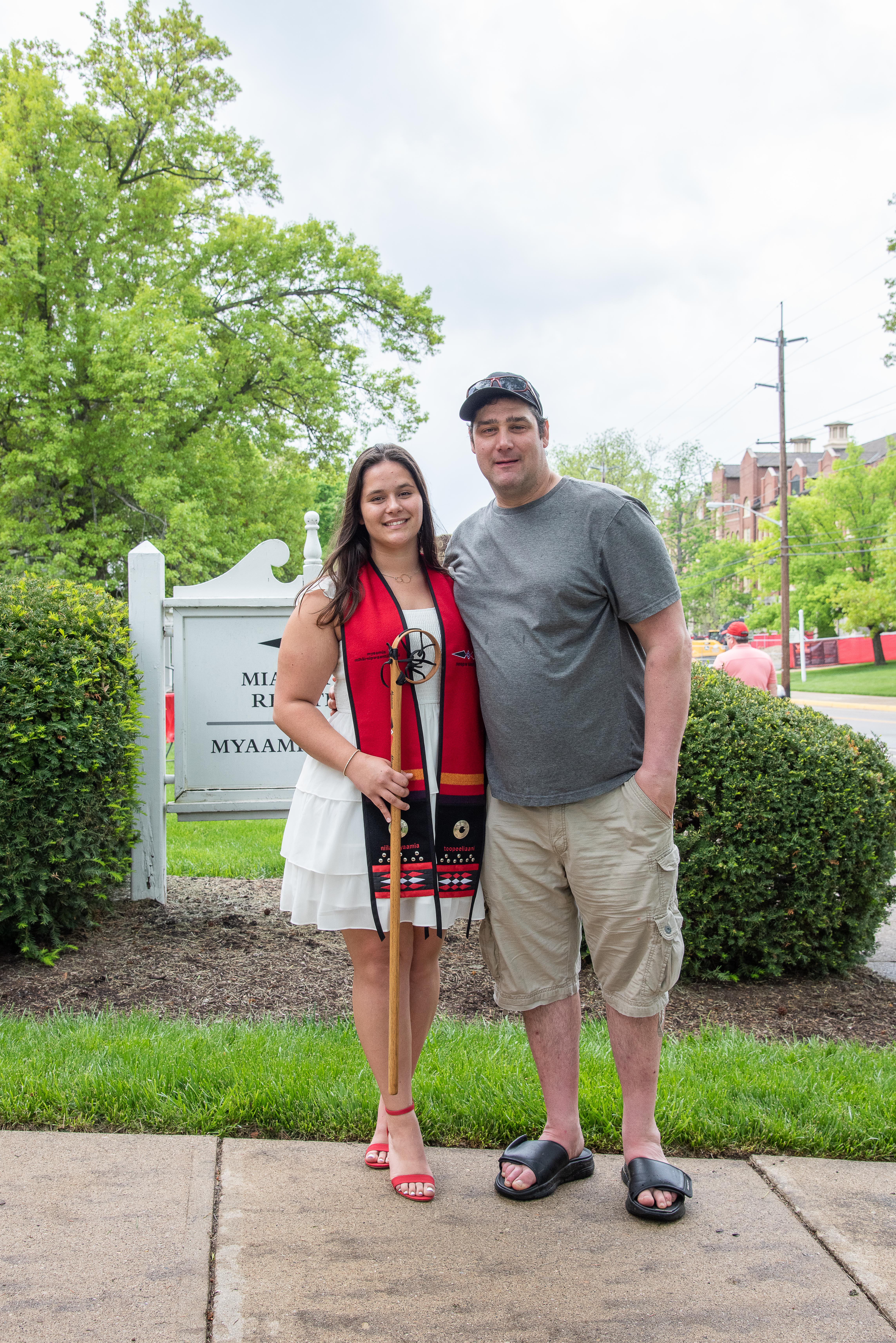 Two people stand outside in front of the Myaamia Center sign. A man has his arm around a graduate holding her lacrosse stick.